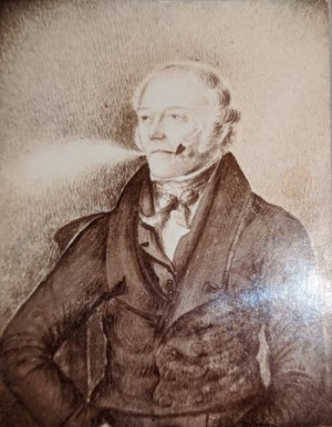 Taylor SWAINSON: a 19th-century gentleman with short, wavy fair or grey hair, clear eyes and a self-possessed air, painted in half profile. He wears a high shirt collar and cravat, a jacket and buttoned-up dark overcoat, and he has a pipe in one corner of his mouth while he blows smoke from the other.